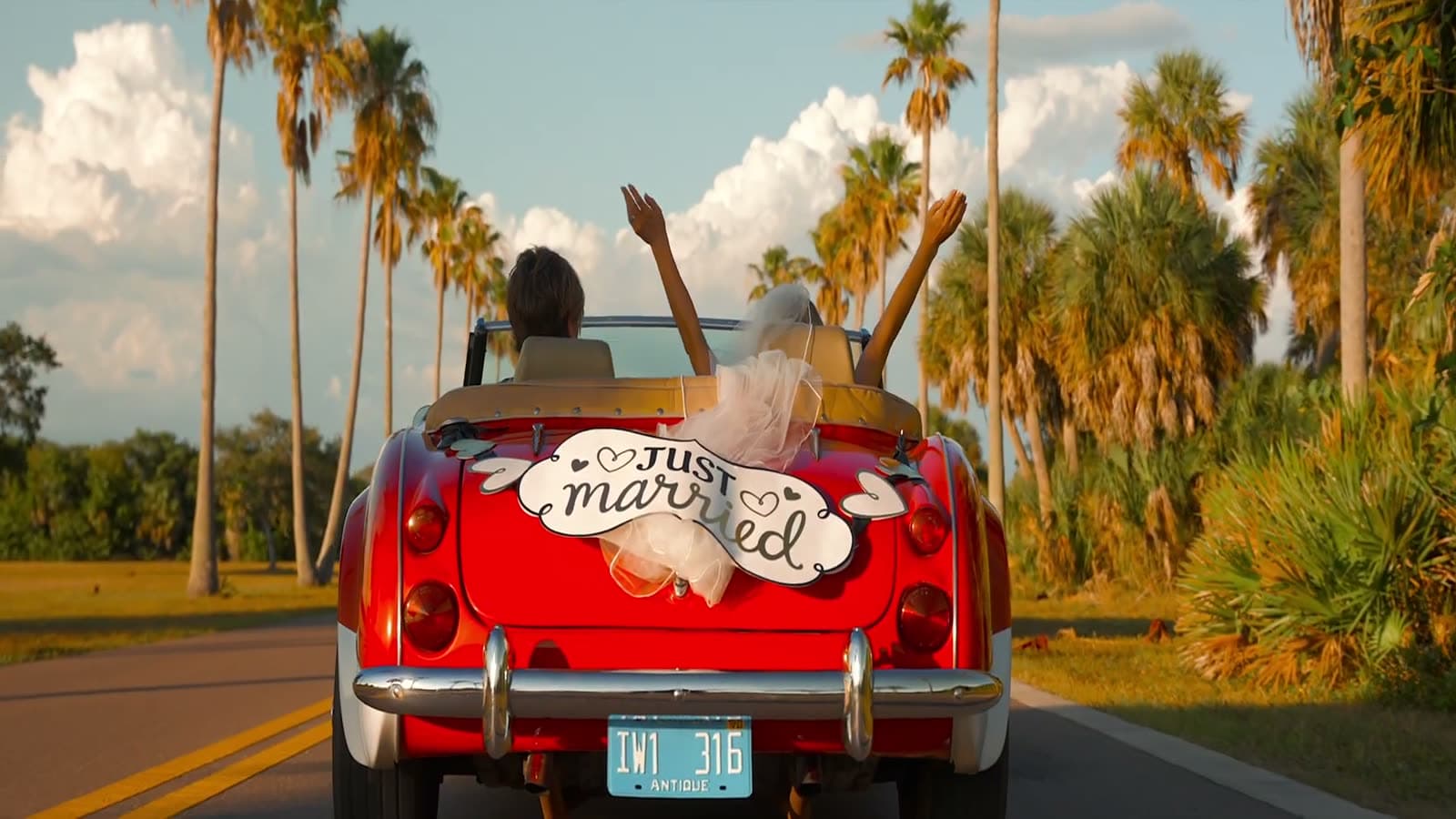 Just-married couple driving a red convertible through palm-lined road, arms raised